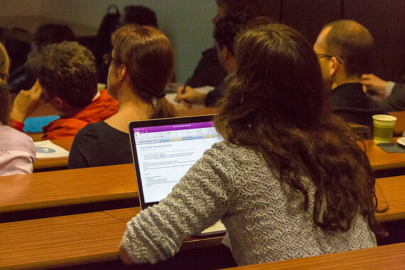 Rows of students, photographed from the back, attending a lecture. One of them with an open laptop.