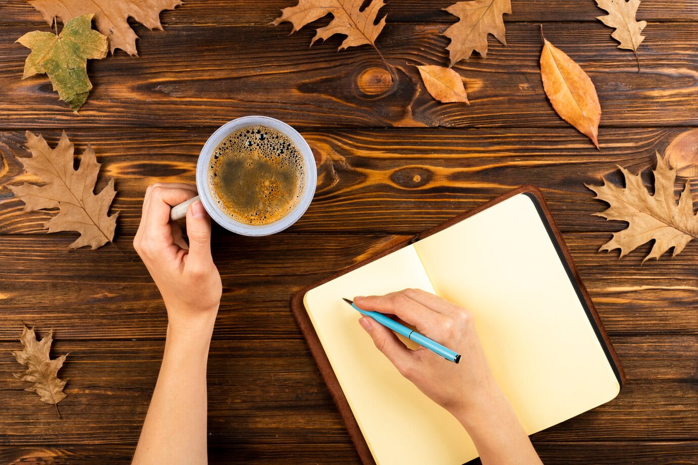 A wooden surface where someone's hands are visible. Left hand is holding a cup of coffee resting on the surface and the right hand is holding a pencil and writing in a blank notebook. Orange autumn leaves seen at the top of the picture.