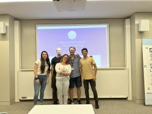 Five organisers of the Distance Based Learning workshop stand in front of a screen, smiling at the camera.