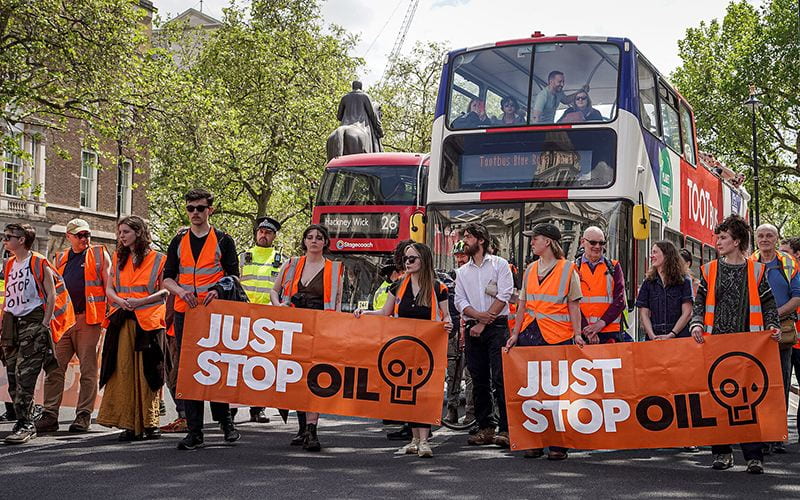 Just Stop Oil protesters with orange banners and hi vis vests protesting along Whitehall, blocking two buses behind them.