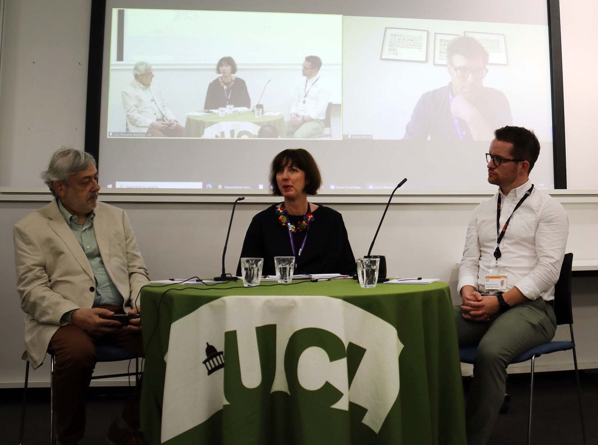 Photo image of three people sat behind a table with the UCL banner on. Behind them is a video screen showing an image of themselves next to a livestream image of a fourth person. 