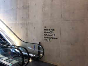 Base of escalator, adjacent to concrete wall with sign up to Level 2 Hub, Library, Refectory and Students' Union UCL
