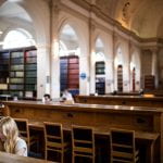 Image by Mat Wright on UCL imagestore. A student with long blonde hair studies in the foreground. Behind her are rows of wooden desks and book stacks in arches sit further back. 