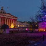 Image by Mary Hinkley on UCL imagestore. UCL front quad at twilight. In front of the portico is a Christmas tree decorated with yellow lights. To the right of the image is a leafless tree decorated with purple and pink lights which can be seen reflecting off the white building beyond. 