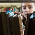 Image by James Tye on UCL imagestore. Image shows a view through a gap in books to a woman with light brown hair holding the books open and appearing to be searching the shelf.