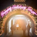 Image by Alejandro Salinas Lopez "alperucho" on UCL imagestore. Image shows a Christmas garland over and arch with people walking through, slightly out of focus. The garland is threaded with yellow lights and the words Happy Holiday Season are written in pink lights.