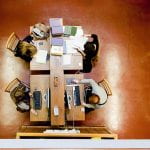 Image by Tony Slade from UCL imagestore. A top-down photograph of four students working individually at wooden desks. To the right of the image are wooden bookcases full of colourful books.