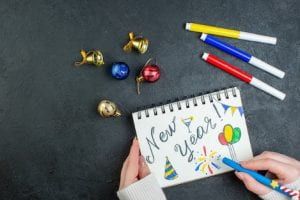 Image by KamranAydinov on Freepik. Top view of hand holding a pen on spiral notebook with new year writing and drawings decoration accessories on black background.