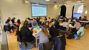 A group of attendees gathered around four rectangular tables engaging in discussions. In the middle of the room, a screen displays the text: "What are the challenges and opportunities that need to be addressed to create equitable conditions in relation to authorship?"