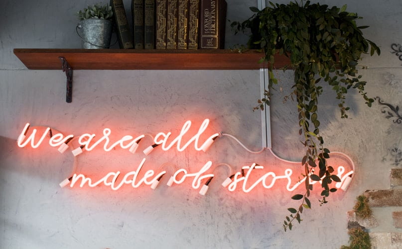 Shelf with books and a plant, above the words 'we are all made of stories' written in red lights