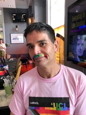 Photograph of Luciano Rila with a rainbow moustache and-shirt with UCL branding in rainbow colours. 
