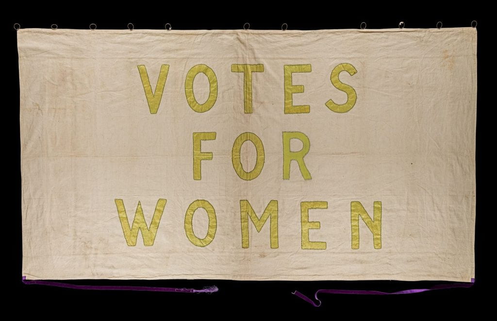 A suffragette banner with white background and green letters stating Votes For Women.