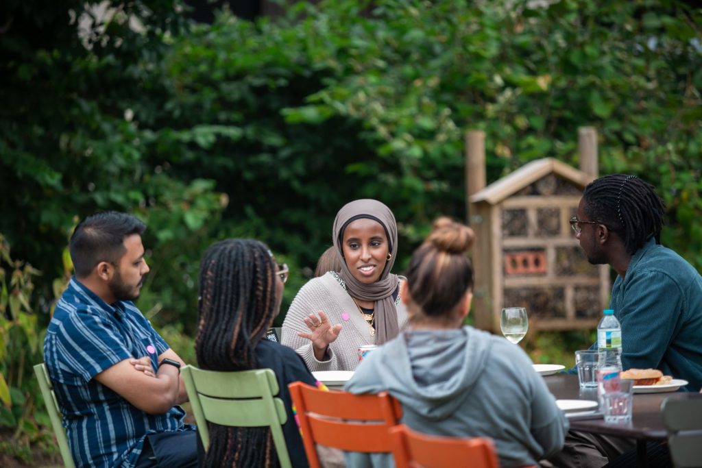 Photo of Salma in a garden sat around the table talking to other young people who's backs are to the camera