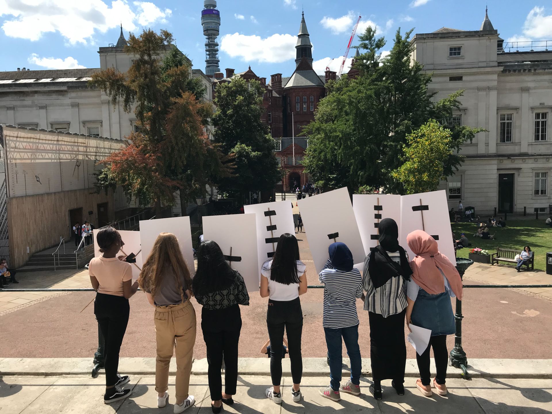 A group of seven Year 12 pupils stand in the UCL main quad holding placards with their backs to the camera.