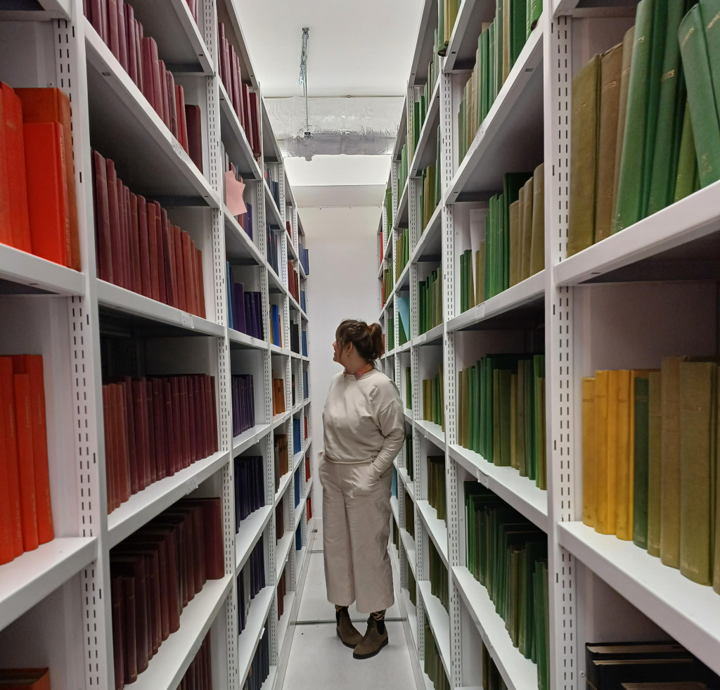 Woman standing between library shelves, looking at a book behind her. 