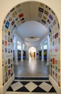 A photograph of an archway leading into the UCL Octagon Gallery. The archway is covered in copies of colourful UCL publication front covers.
