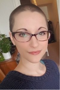 Dr Chloe Farahar photograph: close-up of a white woman with a shaved head, wearing purple glasses, looking into the camera