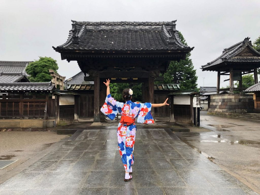 A female student from behind dressed in colourful traditional Japanese clothing striking a pose in front of traditional on a wet day